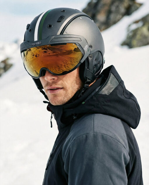 Skifahrer mit dunkelgrauem Casco SP-6 Skihelm mit grün-weißer Zierstreifen und goldfarbenem Visier im verschneiten Gebirge.