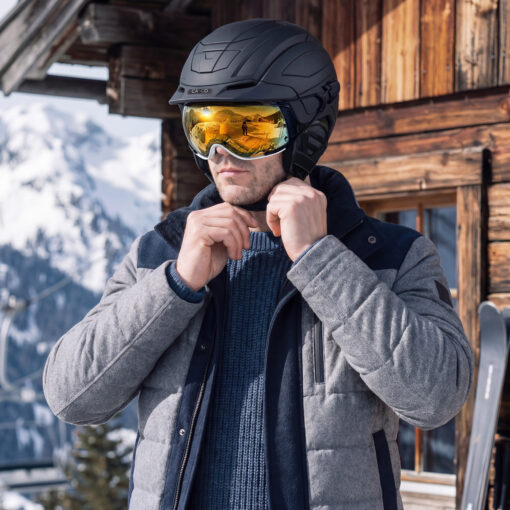 Mann stellt Casco SP-5 Prime Skihelm in Schwarz mit gold verspiegelter Skibrille vor verschneiter Berghütte ein.