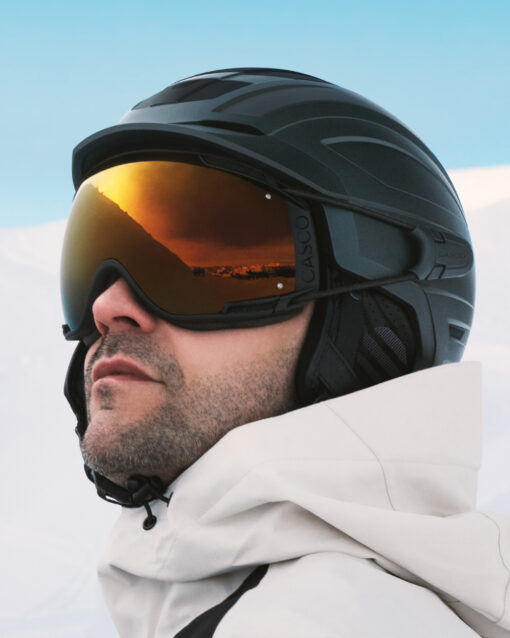 Skifahrer mit schwarzem Casco SP-5 Prime Skihelm und FX-80 Skibrille mit goldverspiegeltem Glas vor winterlicher Bergkulisse.